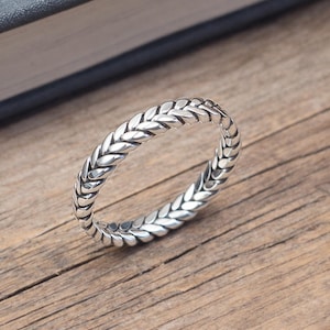 May include: A silver ring with a braided design. The ring is made of a shiny metal and features a repeating pattern of woven links. The ring is displayed on a wooden surface.