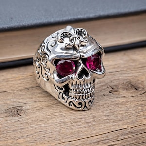 May include: Silver skull ring with intricate detailing and large, red gemstone eyes. The ring has a fleur-de-lis design on the forehead and is set on a wooden surface.