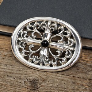 May include: Oval silver-tone belt buckle with an ornate, openwork design. The buckle features a central cross motif with a black stone at the center. The decorative elements include swirling patterns. The buckle is set against a wooden surface.