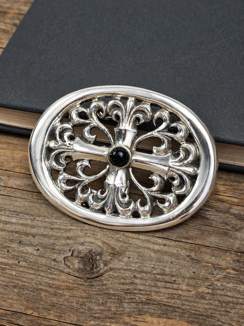 May include: Oval silver-tone belt buckle with an ornate, openwork design. The buckle features a central cross motif with a black stone at the center. The decorative elements include swirling patterns. The buckle is set against a wooden surface.