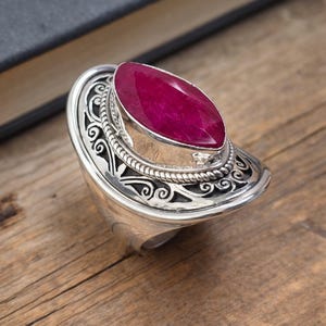 Puede incluir: Anillo de plata con una gema ovalada de color rojo intenso. El anillo presenta un diseño ornamental de filigrana alrededor de la piedra y en la banda. Se encuentra sobre una superficie de madera.