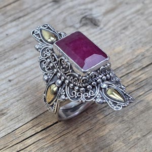 May include: A silver ring with a large, rectangular ruby gemstone. The ring has intricate filigree detailing and gold accents. The ruby is a deep red colour, set against a wooden background.