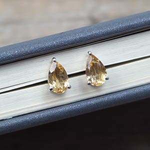 Può includere: Un paio di orecchini a lobo con pietre preziose di citrino a forma di goccia. Le gemme sono di un colore giallo-arancio caldo, incastonate in metallo color argento. Gli orecchini sono appoggiati su un libro con copertina grigia.