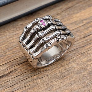 May include: A silver-colored ring featuring a skeletal hand design. The ring has detailed bone structures and a small skull with a pink gemstone. The ring is set on a wooden surface.