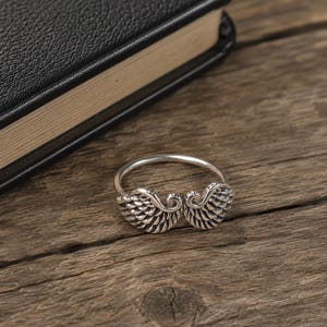 Puede incluir: Un anillo de tono plateado que presenta dos motivos de alas de ángel simétricos y detallados que se encuentran en el centro. El metal tiene un acabado oxidado en las áreas hundidas de las plumas para resaltar la textura y la forma.