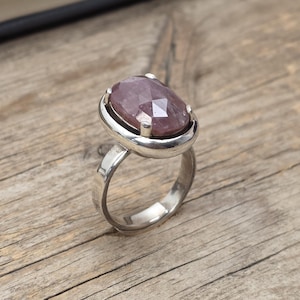 May include: A silver ring featuring a large, oval-cut, pink gemstone. The gemstone is set in a bezel setting, and the ring band is polished silver. The ring is presented on a wooden surface.