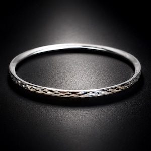 May include: A thin, circular silver bangle bracelet featuring a textured, diamond-cut pattern along its outer surface. The metal has a polished, reflective finish that catches the light, creating a bright, metallic appearance against the dark background.
