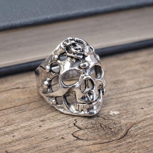 May include: A silver skull ring with intricate detailing, including open eye sockets and a detailed jawline. The ring is set on a wooden surface, with a book in the background. The ring's design is gothic and edgy.