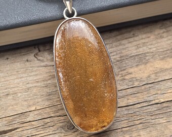 May include: A silver pendant with a large, oval-shaped amber-colored stone. The stone has a textured surface and is set in a silver bezel. The pendant hangs from a silver chain. The background is a wooden surface.
