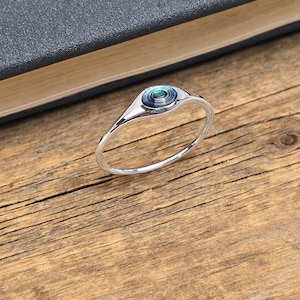 May include: A silver ring featuring a circular, iridescent blue and green gemstone. The ring is placed on a wooden surface beside a dark book. The ring's band is slender and delicate.