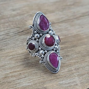 May include: A silver ring featuring four deep red, faceted gemstones. The central stone is round, flanked by two pear-shaped stones at the top and bottom, and a smaller round stone on the side. The metal setting displays intricate beaded and twisted wire details.