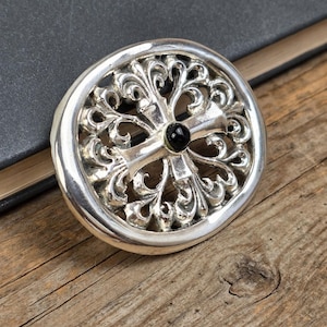 May include: A silver-colored, circular pendant with an ornate, openwork design. The pendant features a cross shape at the center with a black stone. The pendant is resting on a wooden surface.
