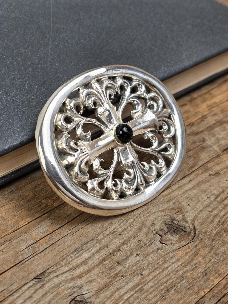 May include: A silver-colored, circular pendant with an ornate, openwork design. The pendant features a cross shape at the center with a black stone. The pendant is resting on a wooden surface.