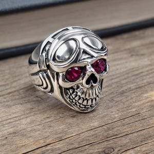 May include: A silver skull ring with ruby-colored eyes. The ring features a detailed design of a skull with goggles. The ring is set on a wooden surface.