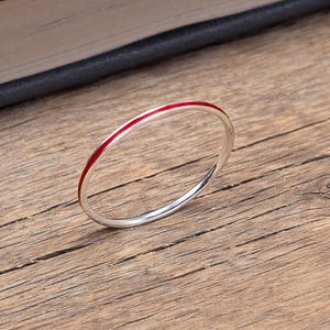 May include: A silver ring with a thin red stripe. The ring is set against a wooden surface, with a book visible in the background. The ring is a simple, elegant piece of jewellery.