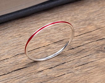 Puede incluir: Un anillo circular fino de plata que presenta una incrustación de esmalte rojo vibrante y liso a lo largo de la circunferencia exterior de la banda. El metal tiene un acabado pulido y reflectante, creando un diseño limpio y sencillo.