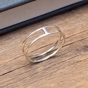 May include: A silver ring with a distinctive design. It has two parallel bands joined by a vertical bar, forming an open, geometric shape. The ring is shown on a wooden surface.