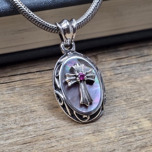 May include: A silver pendant necklace featuring an oval-shaped dark iridescent mother-of-pearl inlay. Centered on the inlay is a silver cross with a small, round, vibrant pink gemstone set at its intersection. The pendant hangs from a silver chain.