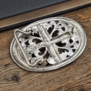 May include: Silver-toned oval belt buckle with an openwork design and a cross in the center. The buckle has a hook and a pin for attachment. The background is a wooden surface.