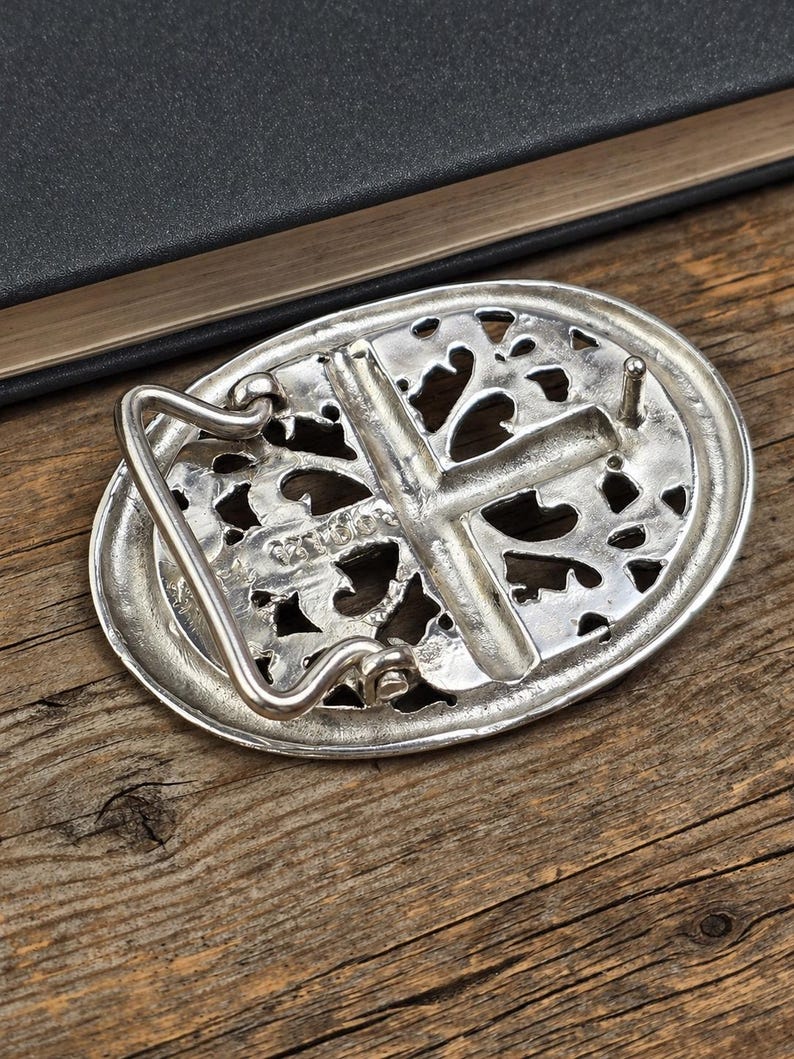 May include: Silver-toned oval belt buckle with an openwork design and a cross in the center. The buckle has a hook and a pin for attachment. The background is a wooden surface.