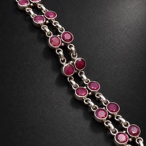 Puede incluir: Una pulsera de doble cadena con rubíes redondos y facetados de color rosa intenso, engastados en biseles de plata pulida. Las piedras están conectadas por eslabones circulares de plata, creando un diseño de cadena continua que resalta las gemas.