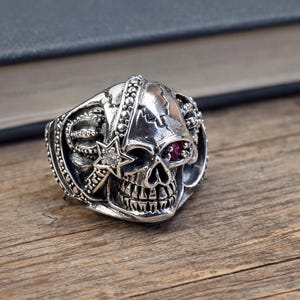 May include: A silver skull ring with red gemstone eyes and a star accent. The ring has intricate detailing and is set against a wooden surface. The skull design is a prominent feature, showcasing a gothic or biker theme.
