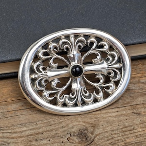 May include: Oval-shaped silver belt buckle with an ornate, openwork design featuring a cross at the center and a black stone. The buckle is set against a wooden surface.