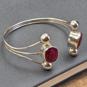 May include: Silver cuff bracelet with three parallel bands. The bracelet features three large, round, deep red gemstones and smaller silver spheres. The bracelet is resting on a dark grey surface.