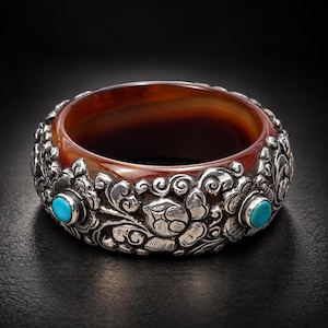 May include: A wide bangle bracelet featuring a translucent deep reddish-brown stone band encased in intricate silver metalwork. The silver overlay displays embossed floral patterns and is accented with small, round, bright blue turquoise stones.