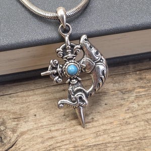 May include: A silver pendant necklace with an ornate, stylized design. The pendant features a central turquoise-colored stone and intricate details. The necklace is displayed on a wooden surface.