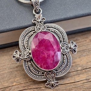 May include: A pendant necklace featuring a large, oval-shaped, faceted magenta gemstone set in an intricate sterling silver frame. The silver setting displays detailed filigree work, beaded textures, and four symmetrical floral-like accents.