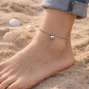 May include: A silver-toned anklet featuring a delicate chain of small metallic beads and a single, polished heart-shaped charm suspended from the center. The jewelry piece rests around the ankle, showcasing a simple and reflective metallic finish.