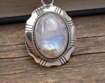 May include: A silver pendant necklace featuring an oval, iridescent moonstone. The stone is set in a scalloped, textured silver frame with a rope-like border. The necklace rests on a wooden surface.