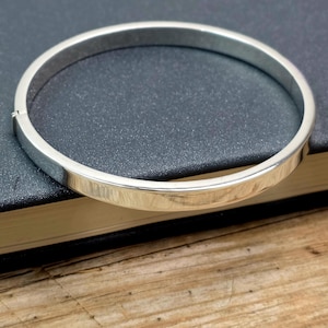 May include: A silver bangle bracelet with a flat, polished surface. The bracelet is circular and has a simple, elegant design. The jewellery is shown on a dark surface, with a wooden surface in the background.