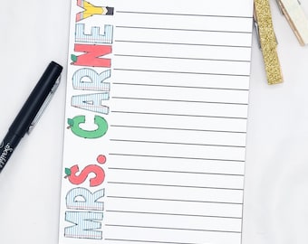 May include: A lined notepad with the text "Mrs. Carney" in colorful, playful lettering. A black pen rests on the left, and two decorative clothespins are on the right. The notepad is white with black lines.