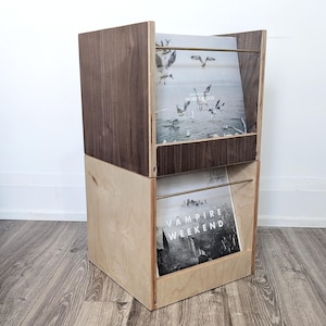 May include: A two-tiered wooden record storage unit with a dark brown stain on the top tier and a natural wood finish on the bottom tier. The unit is holding two vinyl records, one with the text "Dan Deacon More or Less" and the other with the text "Vampire Weekend".