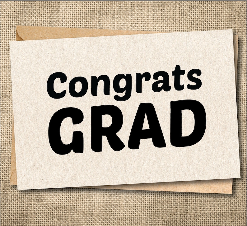 Peut inclure: Une carte de f&eacute;licitations de fin d'&eacute;tudes avec les mots "Congrats GRAD" en lettres noires en gras. La carte est beige clair et repose sur une enveloppe marron. Le fond est un tissu textur&eacute;, beige.
