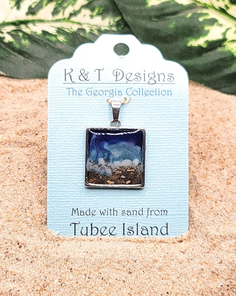 Op de afbeelding: Een zilveren ketting met een vierkante hanger van hars met blauw, wit en bruin zand van Tubbee Island. Het label luidt "K&T Designs The Georgia Collection Made with sand from Tubbee Island."