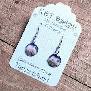 Op de afbeelding: Een paar zilveren oorbellen met kleine ronde blauwe en witte harsbedels die zand van Tybee Island bevatten. De oorbellen bevinden zich op een witte kaart met de tekst "K&T Designs The Shoreline Collection Made with sand from Tybee Island".