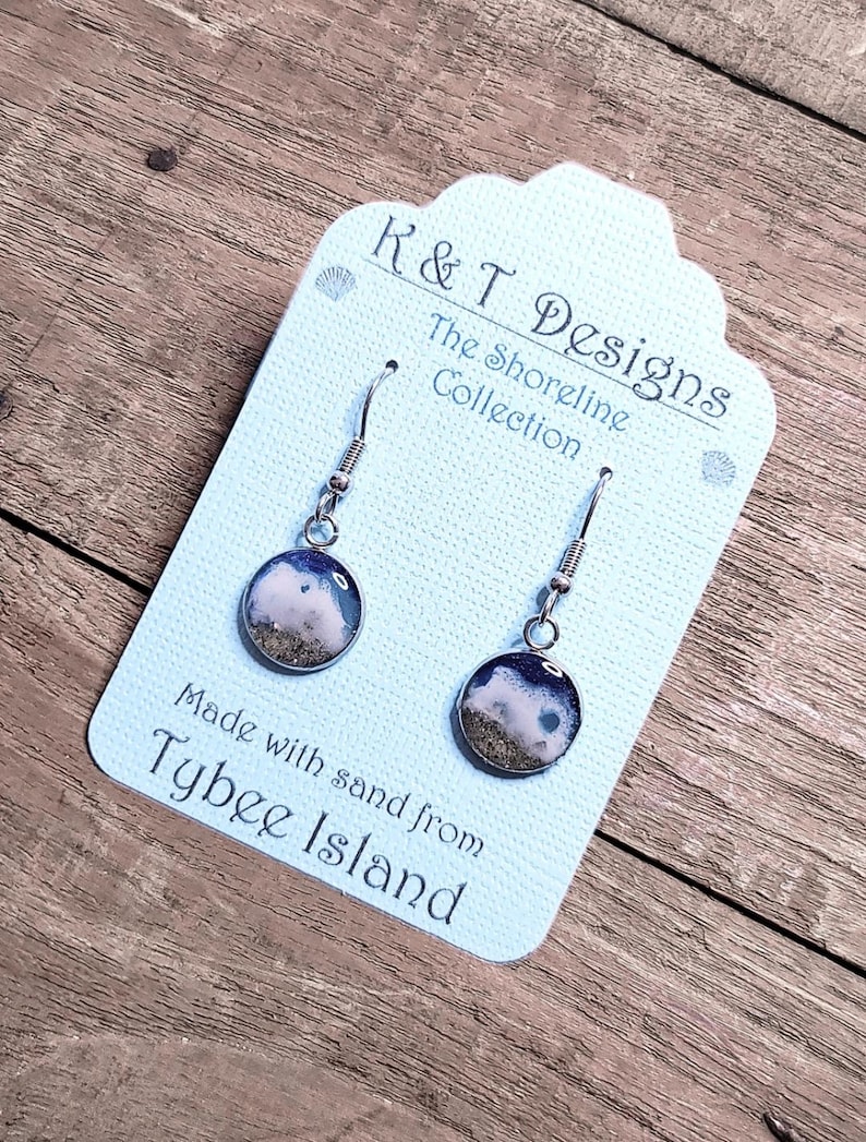 Op de afbeelding: Een paar zilveren oorbellen met kleine ronde blauwe en witte harsbedels die zand van Tybee Island bevatten. De oorbellen bevinden zich op een witte kaart met de tekst "K&T Designs The Shoreline Collection Made with sand from Tybee Island".