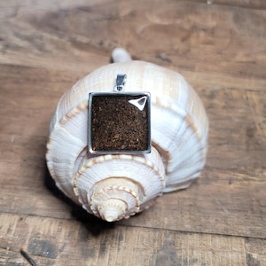 May include: A square pendant with a silver frame and a brown, glittery resin insert. The pendant is sitting on a white seashell.