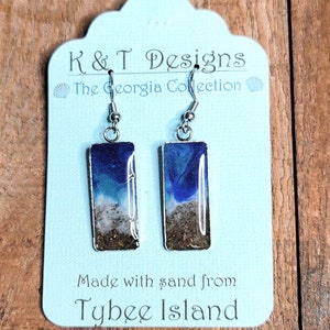 Op de afbeelding: Een paar zilveren oorhangers met blauwe, witte en bruine harsinzetstukken. De hars is gemaakt met zand van Tybee Island. De oorbellen bevinden zich op een blauwe en witte kaart met de tekst "K & T Designs The Georgia Collection" en "Made with sand from Tybee Island."