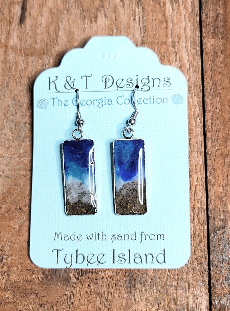 Op de afbeelding: Een paar zilveren oorhangers met blauwe, witte en bruine harsinzetstukken. De hars is gemaakt met zand van Tybee Island. De oorbellen bevinden zich op een blauwe en witte kaart met de tekst "K & T Designs The Georgia Collection" en "Made with sand from Tybee Island."
