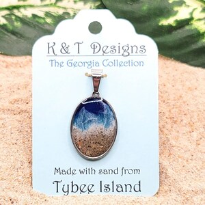 Op de afbeelding: Een zilveren ketting met een hanger van helder, ovaal hars, met zand van Tybee Island, Georgia. De hanger bevindt zich op een wit label met de tekst "K & T Designs The Georgia Collection Made with sand from Tybee Island".