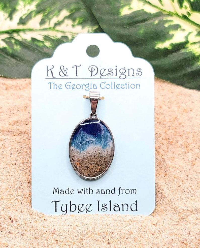 Op de afbeelding: Een zilveren ketting met een hanger van helder, ovaal hars, met zand van Tybee Island, Georgia. De hanger bevindt zich op een wit label met de tekst "K & T Designs The Georgia Collection Made with sand from Tybee Island".