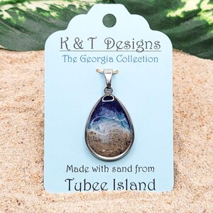 Op de afbeelding: Een traanvormige hangerketting met een zilveren ketting. De hanger is gevuld met zand en heeft een blauw en wit golfontwerp. Het label luidt "K & T Designs The Georgia Collection Made with sand from Tybee Island".