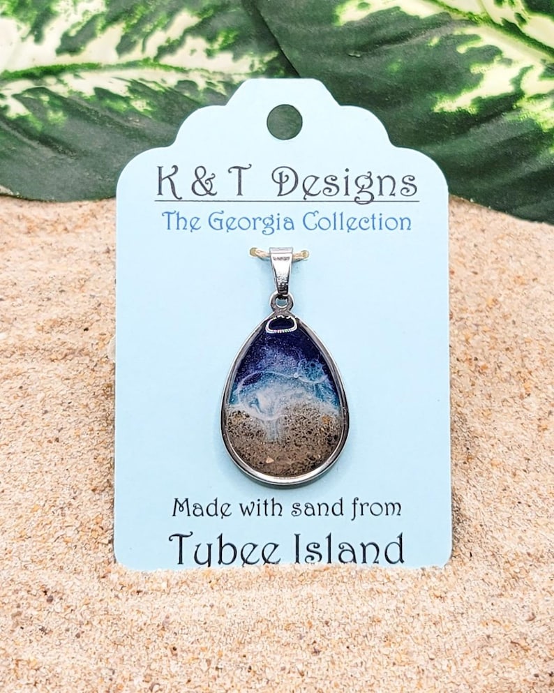 Op de afbeelding: Een traanvormige hangerketting met een zilveren ketting. De hanger is gevuld met zand en heeft een blauw en wit golfontwerp. Het label luidt "K & T Designs The Georgia Collection Made with sand from Tybee Island".