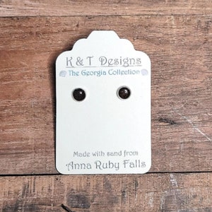 May include: A pair of brown stud earrings made with sand from Anna Ruby Falls. The earrings are displayed on a white card with the text "K & T Designs The Georgia Collection" and "Made with sand from Anna Ruby Falls".