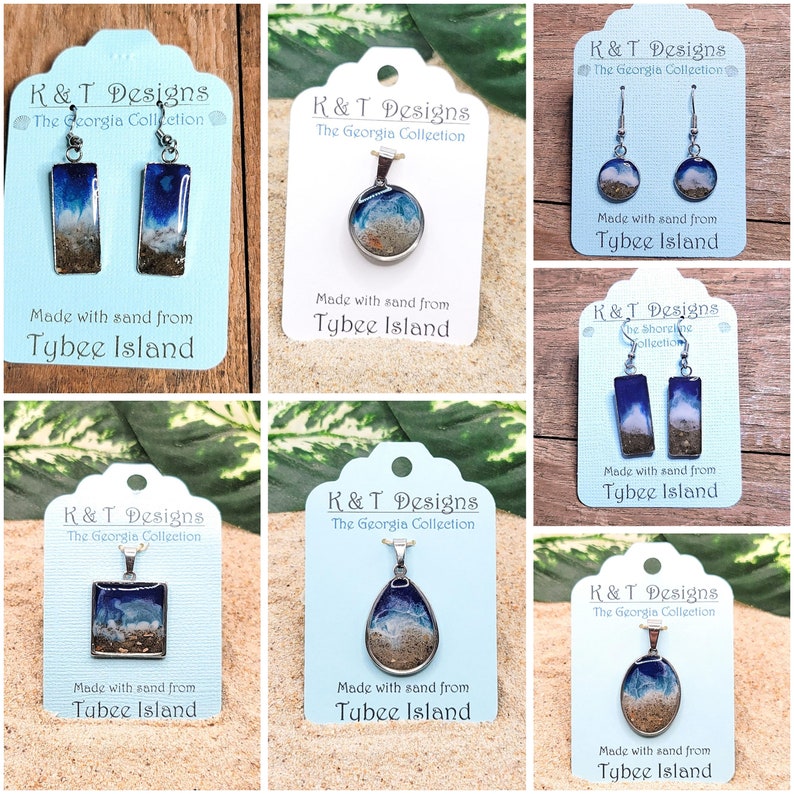 Op de afbeelding: Zes verschillende sieraden met Tybee Island-zand ingekapseld in hars. Elk stuk wordt getoond op een witte kaart met de tekst "K&T Designs The Georgia Collection" en "Gemaakt met zand van Tybee Island". De sieraden omvatten oorbellen, hangers en kettingen.