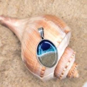 May include: A silver pendant with a blue and white resin inlay that resembles a beach scene. The pendant is oval shaped and has a silver bail.